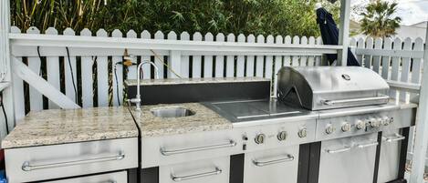 Une cuisine extérieure en acier inoxydable, dotée d’un évier, d’un barbecue intégré et d’une clôture blanche en bois en arrière-plan.