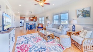 A spacious living area with a large sofa, a coffee table, and a dining table with chairs. There is a ceiling fan, a television, and artwork on the walls. - Oceanfront Condo w/ Private Sundeck, Resort Pool, Elevator, and Beach Access (Rodanthe)