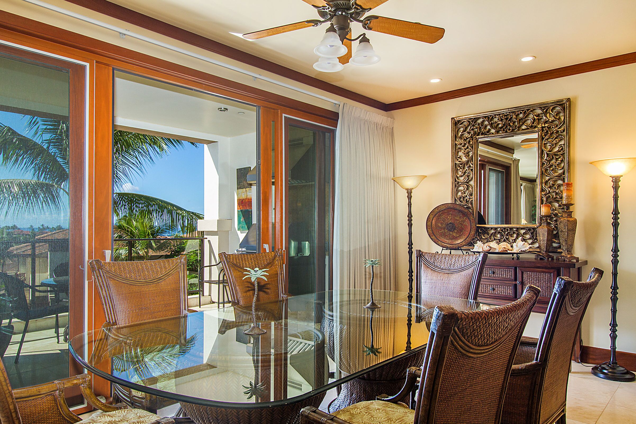 A dining area with a glass table and wicker chairs, a ceiling fan, and a large mirror on the wall.