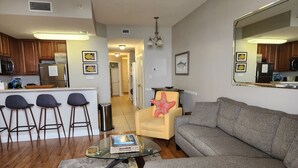 A modern living room with a grey sofa, a glass coffee table, and a starfish cushion. There are wooden chairs and a kitchen in the background.