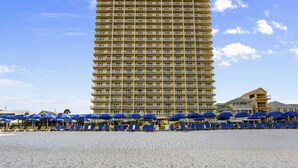 A beachfront hotel with a tall building, multiple balconies, and blue umbrellas on the sand.