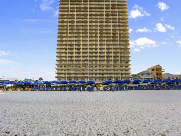 A beachfront hotel with a tall building, multiple balconies, and blue umbrellas on the sand.