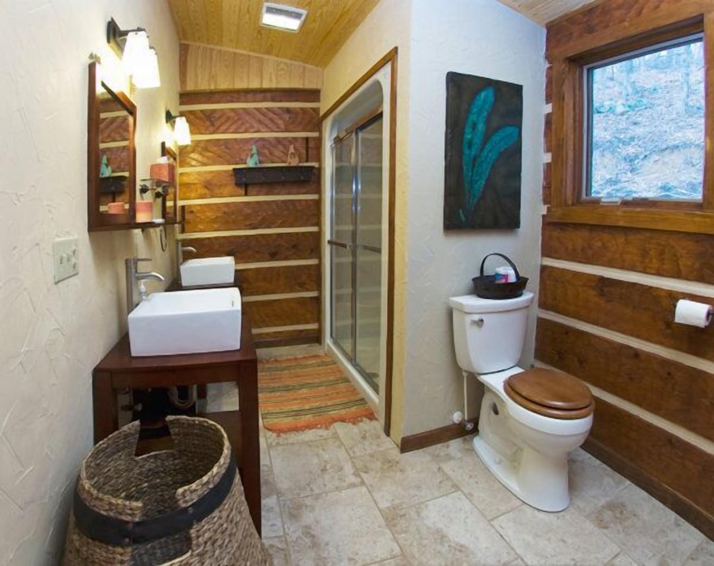 Un baño con detalles de madera, un lavamanos blanco, un inodoro con asiento de madera, una puerta de ducha de vidrio y una ventana con vista a árboles.