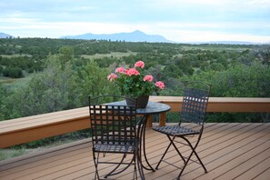 Una terraza de madera con dos sillas de metal y una mesa, con vistas a un paisaje exuberante con árboles y montañas a lo lejos.