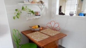 Een kleine keuken met een houten tafel, een groene stoel, een mand met fruit en een decoratieve rode bloem aan de muur.