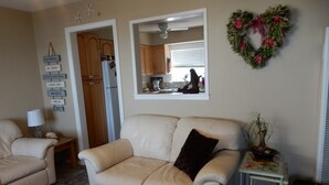 Een woonkamer met een beige bank, een klein tafeltje met een lamp, een decoratieve muur met motiverende bordjes, een keuken zichtbaar door een open deur, en een krans aan de muur.