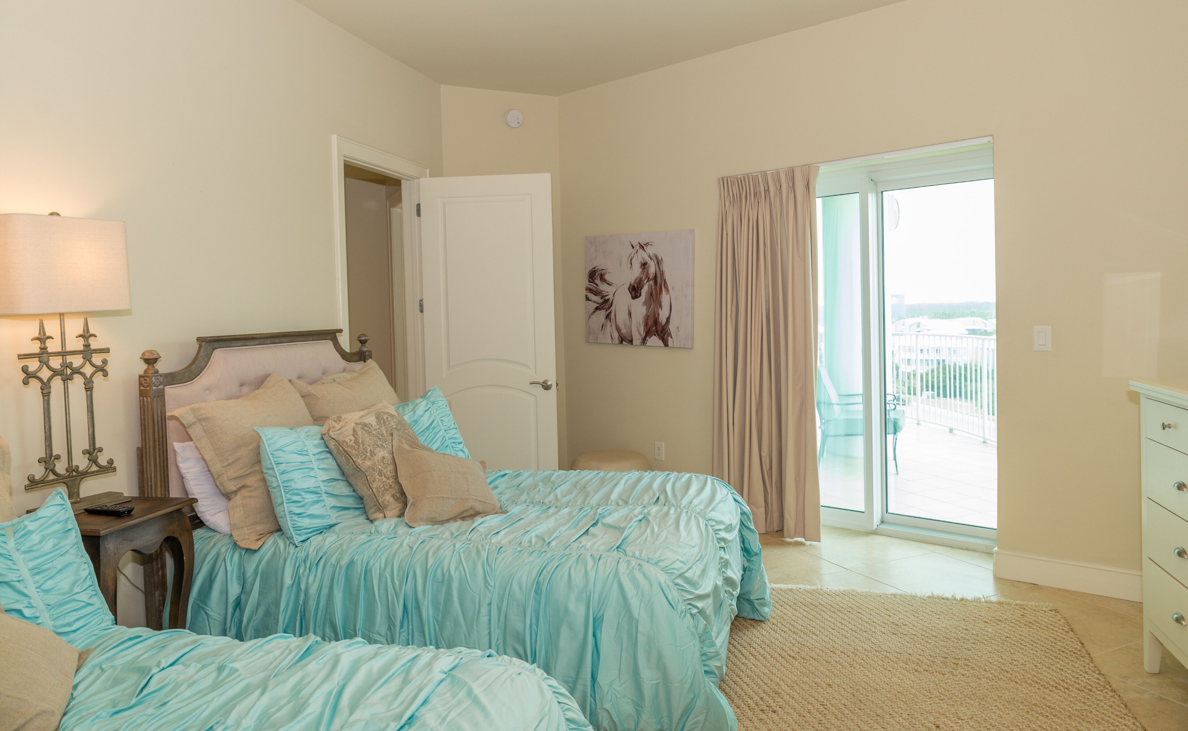 A bedroom with two beds, a nightstand, a lamp, a painting, and a view of the outside.