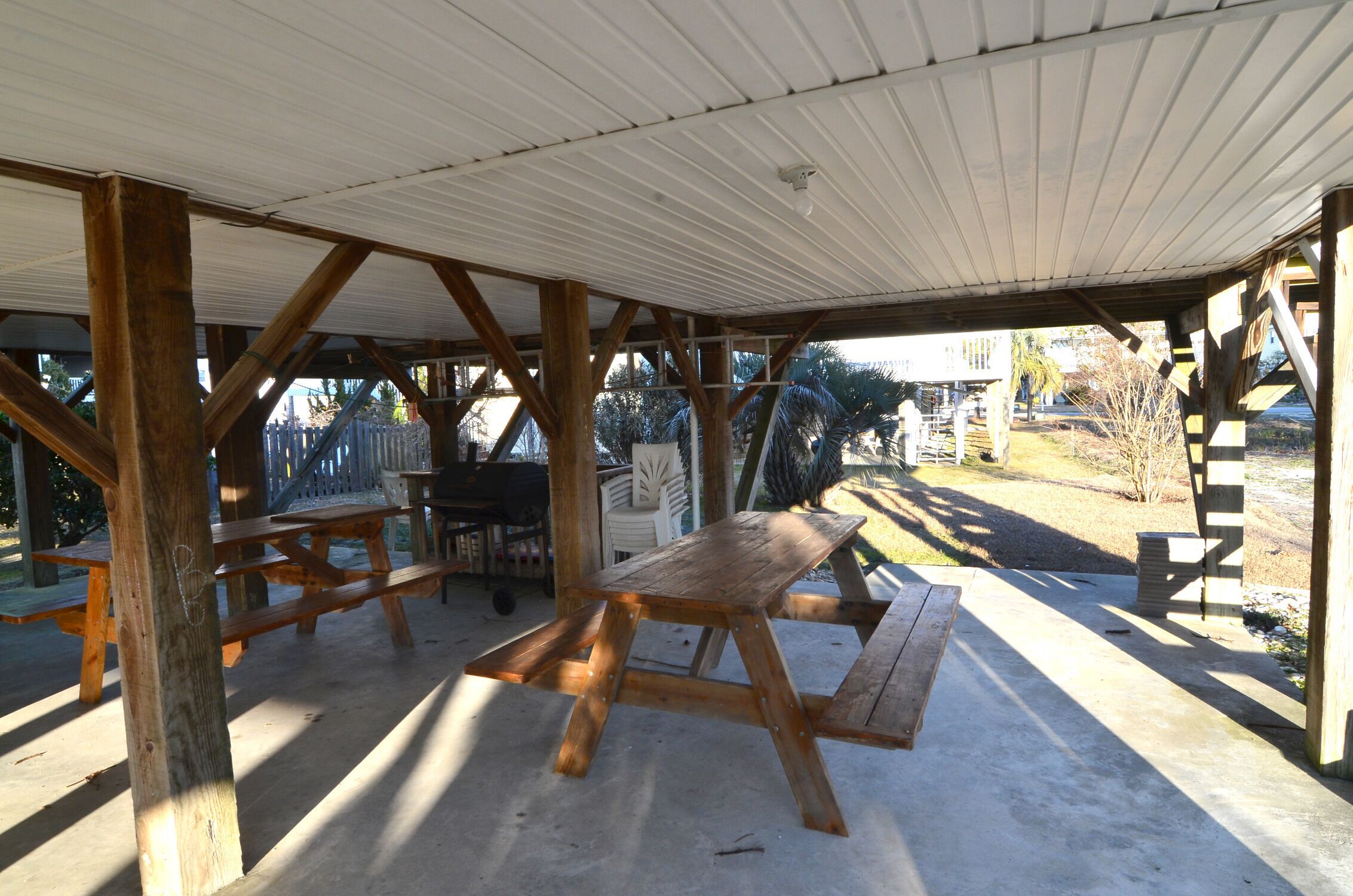 A covered picnic area with wooden benches and tables, surrounded by a wooden fence and palm trees.