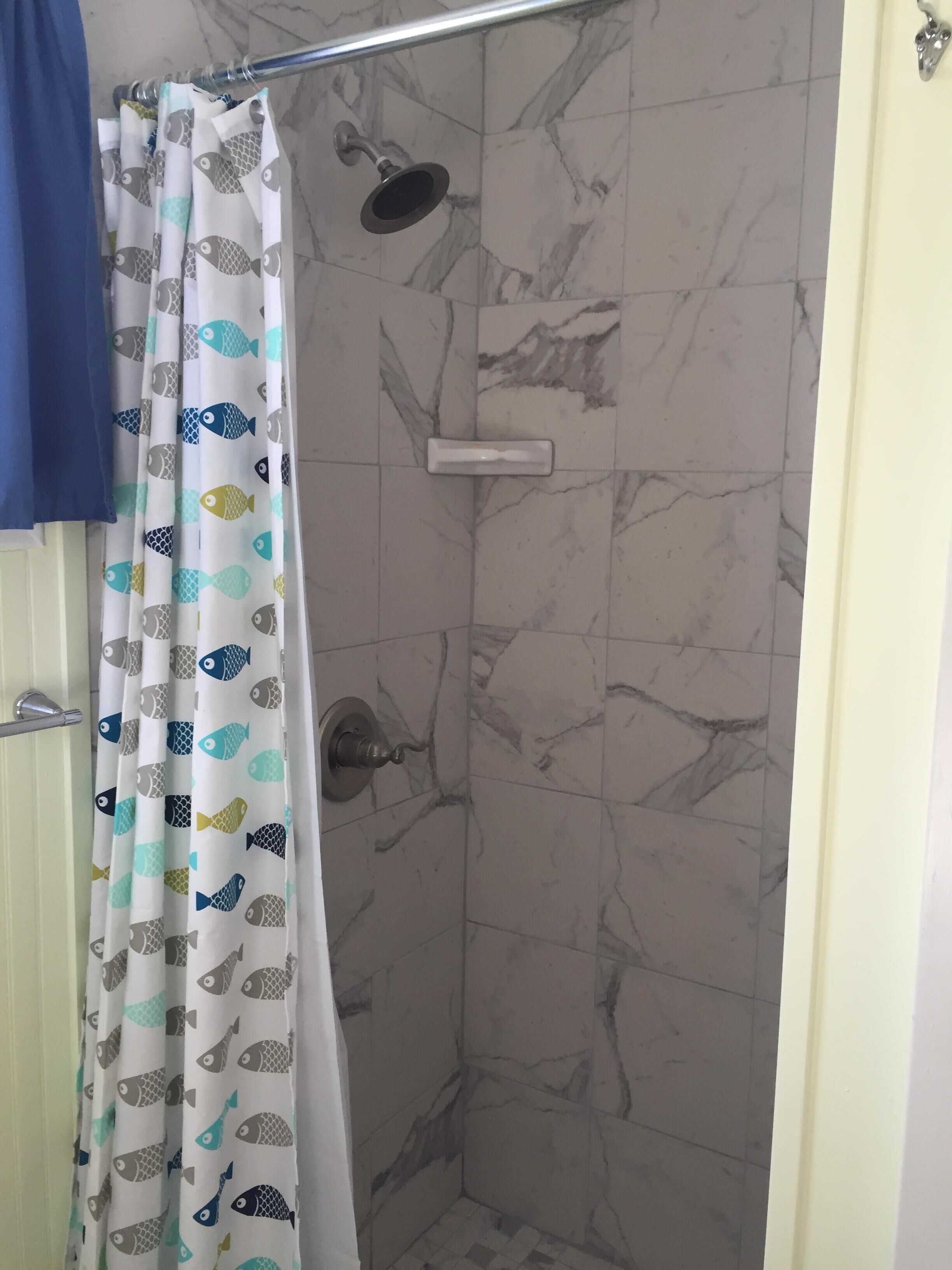 A shower with a fish-patterned curtain, marble tiles, and a handheld showerhead.