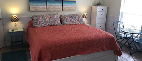 A bedroom with a bed, patterned pillows, a nightstand with a lamp, a dresser, and a window with a view.