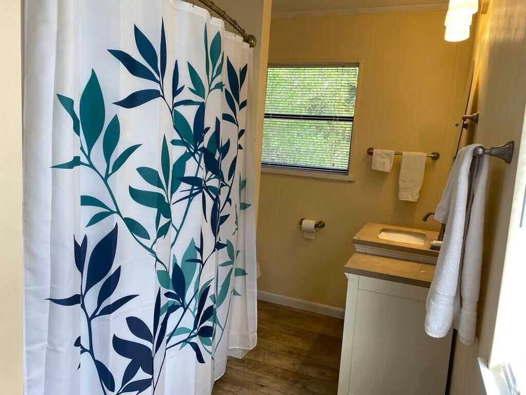 Une salle de bain avec un rideau de douche orné d’un motif de feuilles, une fenêtre avec des stores, un porte-serviettes et un lavabo.