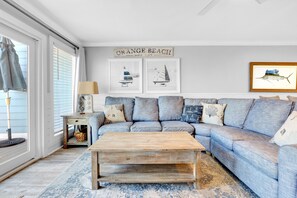 En stue med en blå sofa, et træbord og et tæppe. To billeder hænger på væggen, og et skilt siger "ORANGE BEACH".