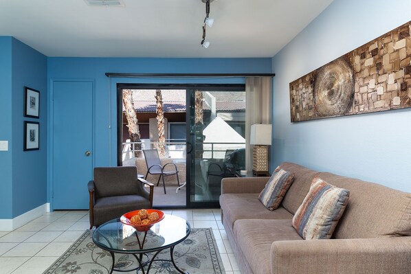 Una sala de estar con una pared azul, un sofá marrón, una mesa de centro de vidrio y una silla. Hay una puerta corrediza de vidrio que da a un área exterior con una silla y una mesa.