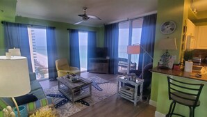 A living room with a sofa, coffee table, and a TV. There is a balcony with a view of a building.