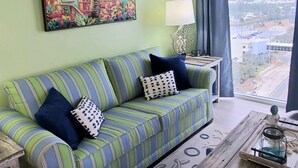 A living room with a green sofa, striped cushions, a wooden coffee table, and a view of a cityscape through a large window.