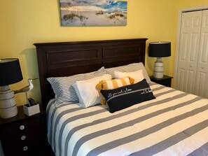 Una cama con ropa de cama a rayas, una mesita de noche con una lámpara, un buró con lámpara y un cuadro en la pared.