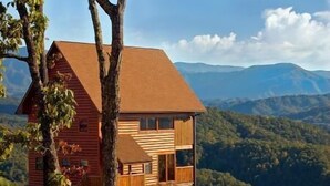 A two-story log cabin with a sloped roof, surrounded by a forested landscape and mountains in the background. - Mountain Splendor: Best Mountain View in Starr Crest Resort - Brand New Hot Tub (Sevierville)