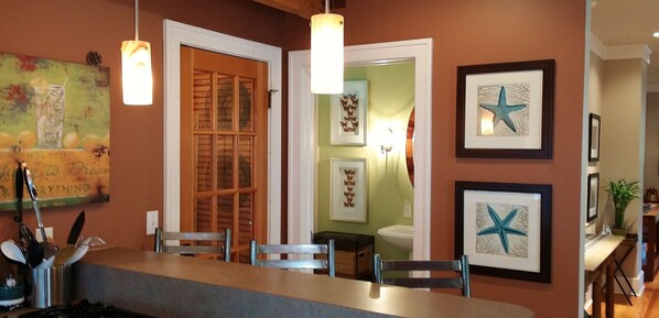 A kitchen with a bar area, wooden cabinets, and a dining table with chairs. There are framed artworks on the walls and a hanging light fixture above the bar.
