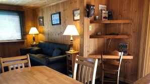 A wooden interior with a round wooden table, two chairs, a sofa, and a bench. There are two wall-mounted lamps, a shelf with items, and framed pictures on the wall. - Black River Retreat-Dog Friendly! (Bessemer)