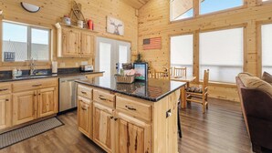 A wooden cabin interior with a kitchen, dining area, and a large window.