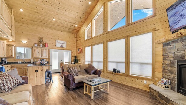 A spacious living area with a wooden ceiling, large windows, a stone fireplace, and a cozy seating arrangement.