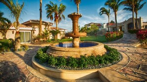 Une fontaine circulaire en pierre, à plusieurs niveaux, entourée d’un jardin aménagé avec des palmiers et surplombée d’un ciel bleu azur.