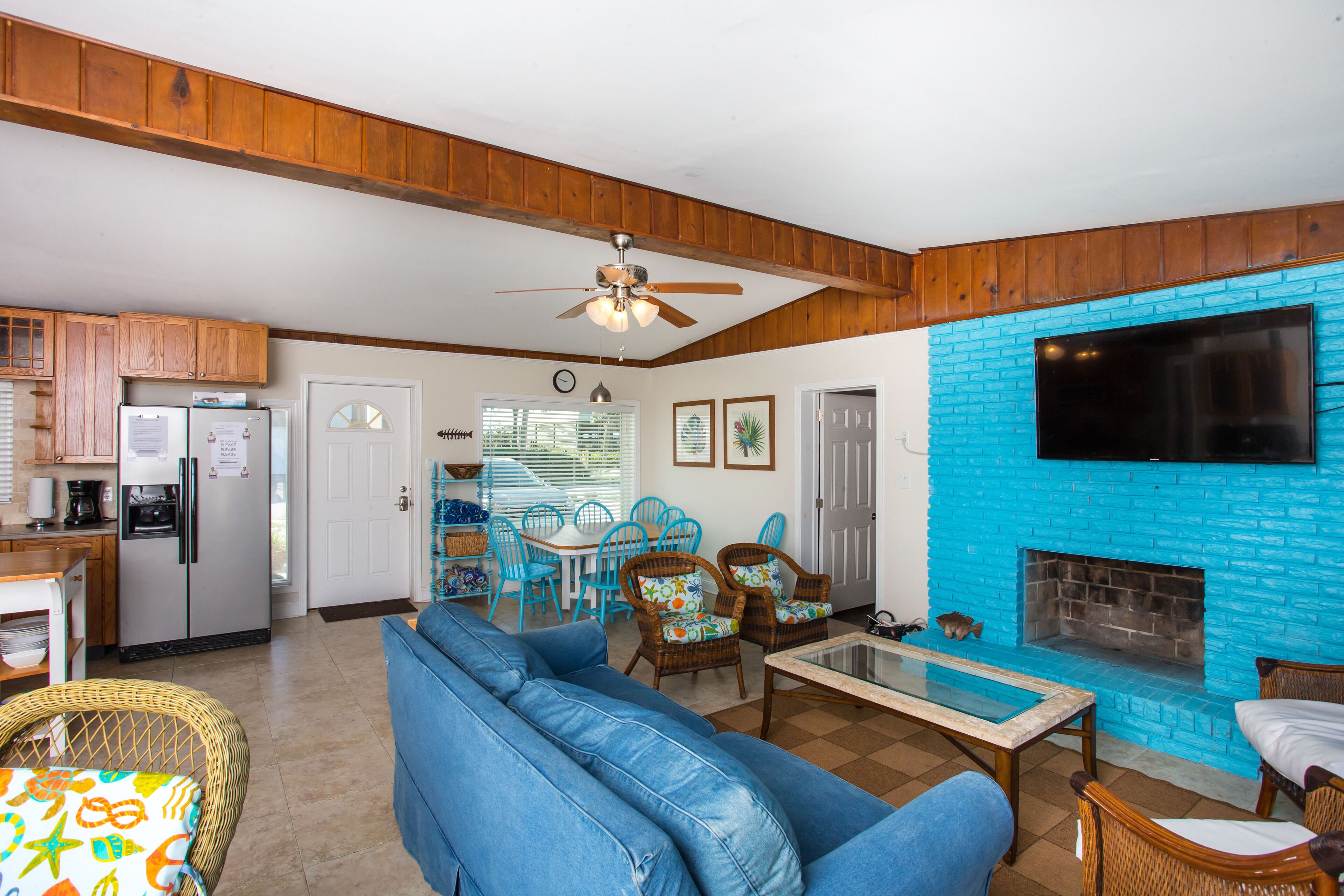 A living room with a blue brick fireplace, a ceiling fan, and a dining area with blue chairs.