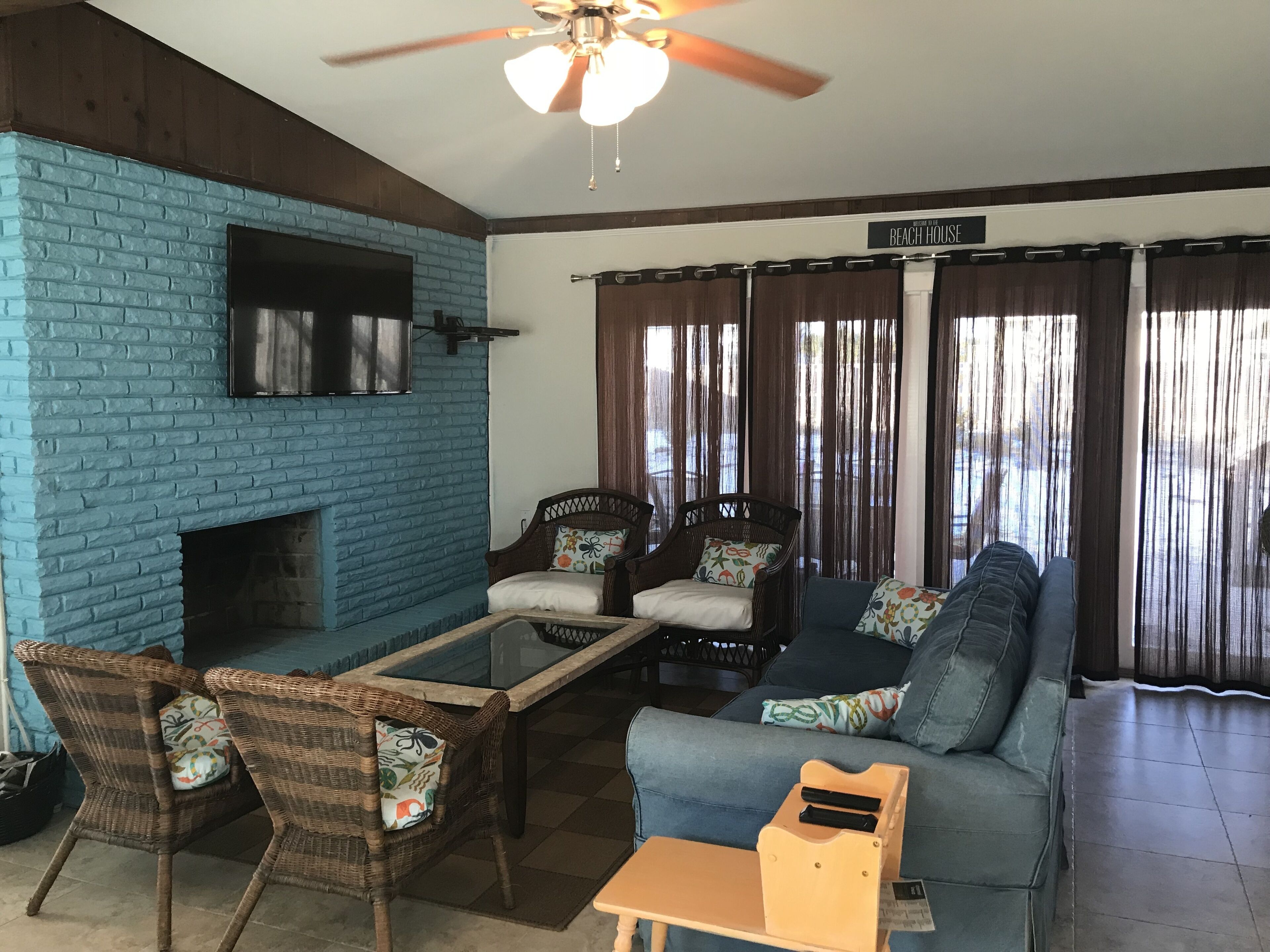 Una sala de estar con pared de ladrillo azul, muebles de mimbre, un ventilador de techo y un letrero que dice "Beach House".