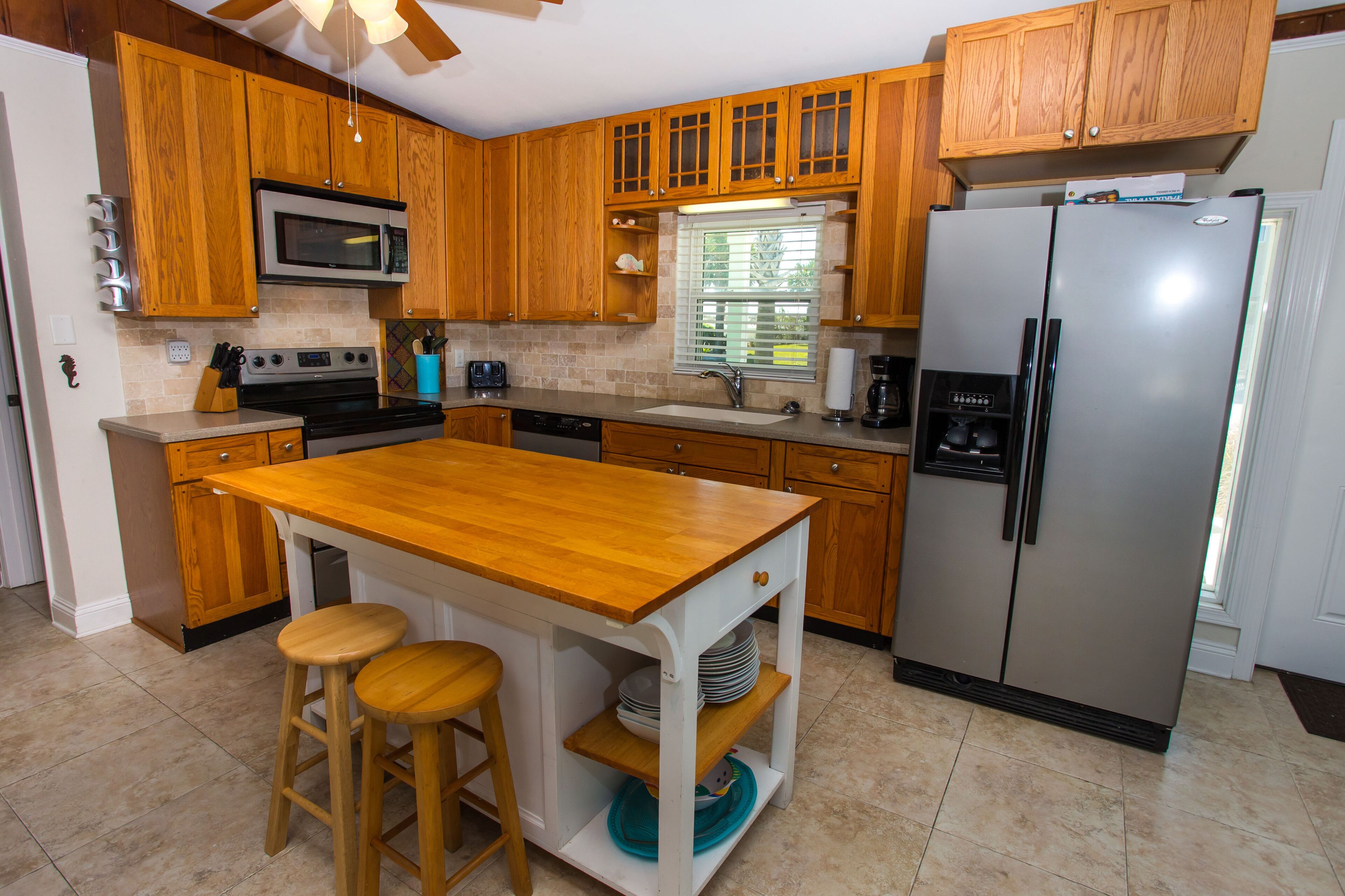 A kitchen with wooden cabinets, a stainless steel refrigerator, a microwave, a sink, and a wooden island with stools.