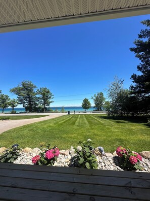 A view from a covered deck overlooking a well-maintained lawn with a gravel border and flowers, a clear blue sky, and a distant body of water. - Lake Michigan Waterfront Retreat - Traverse City, MI (Traverse City)