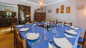 Een eetkamer met een blauwe tafelkleed, houten stoelen en een kroonluchter. Er is een houten kast met glazen en een wijnfles op de tafel.