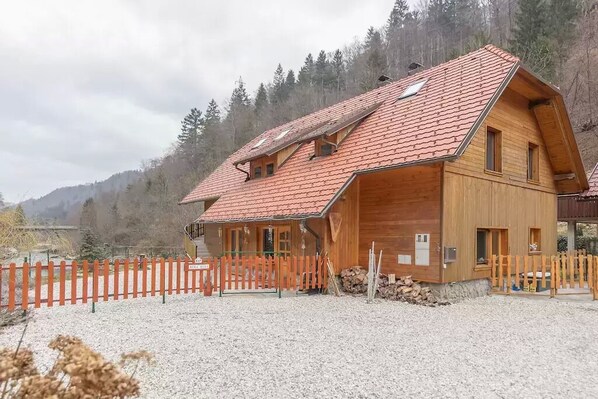 A wooden chalet with a sloped red-tiled roof, a wooden porch, and a gravel driveway. - Reka Hisa River Side Chalet (Bled)