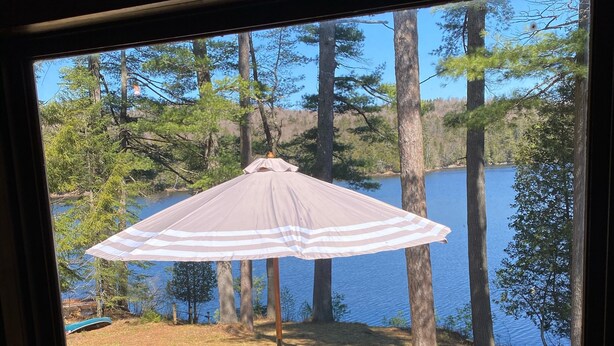 Une vue depuis une fenêtre donnant sur une terrasse avec une table et des chaises, surplombant un lac entouré d’arbres.