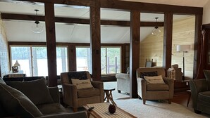 Un salon spacieux avec un plafond haut, des poutres apparentes et de grandes fenêtres offrant une vue sur l’extérieur.
