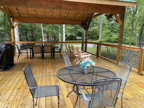 Une terrasse en bois dotée d’un espace salon couvert, de tables et de chaises, le tout entouré d’une balustrade en bois et d’arbres.