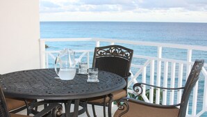 Una mesa metálica redonda con una jarrón y vasos, una silla de mimbre y una barandilla blanca con vistas al mar.