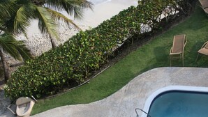 Una piscina con borde de hormigón, una zona ajardinada con seto y una playa al fondo.