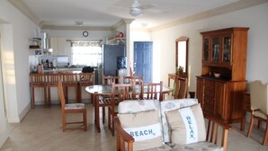 Amplia sala de estar con una mesa de comedor rodeada de sillas de madera, un sofá con cojines de temática playera y un mueble de madera.