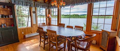 Comedor de madera con una mesa grande y sillas, ventanas amplias con vista a un lago y un candelabro.