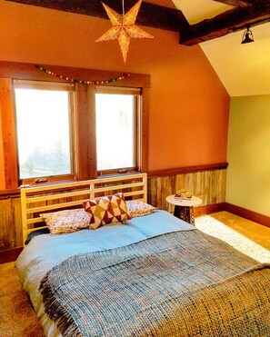 Une chambre confortable avec un lit en bois, une décoration suspendue en forme d’étoile, une fenêtre avec des rideaux et une petite table ronde.