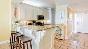 A kitchen with white cabinetry, a granite countertop, and a built-in microwave. There are two wooden stools and a small island with a sink.