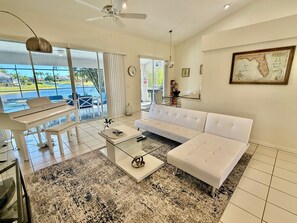 A modern living room with a white sofa, a glass coffee table, and a view of a pool through large windows.