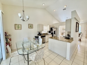 A modern kitchen with a dining area, featuring a glass table, white cabinetry, and a chandelier.
