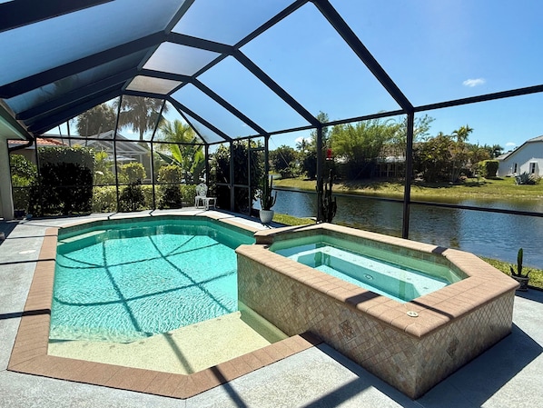 A pool area with a glass-enclosed structure, a hot tub, and a view of a body of water and greenery.