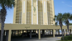 Um hotel com vários andares, entrada central, estacionamento subterrâneo e palmeiras.
