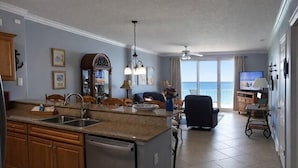 Cozinha equipada com máquina de lavar louça, sala de jantar com mesa e cadeiras, e vista para o mar através de amplas janelas.