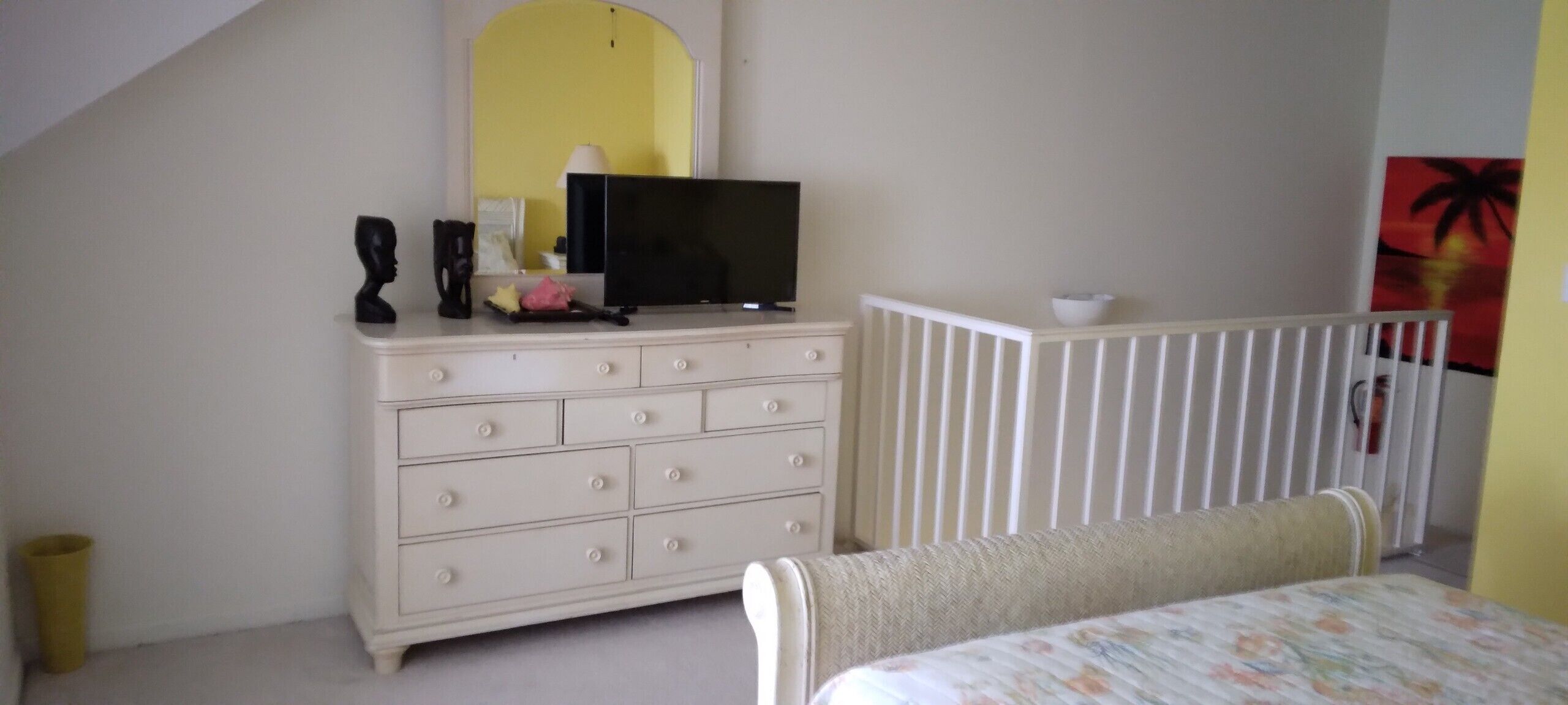 Quarto com cômoda branca, televisão, berço e cama com colcha florida.