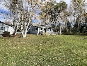 A single-story house with a porch, surrounded by a grassy yard and trees. - Quaint Lakefront Escape, private shoreline, Dock, Kayaks, Sleeps 6 Near Petoskey (Alanson)