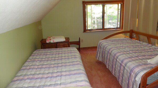 A bedroom with two single beds, a wooden headboard, a nightstand with a lamp, and a window with a view of trees.