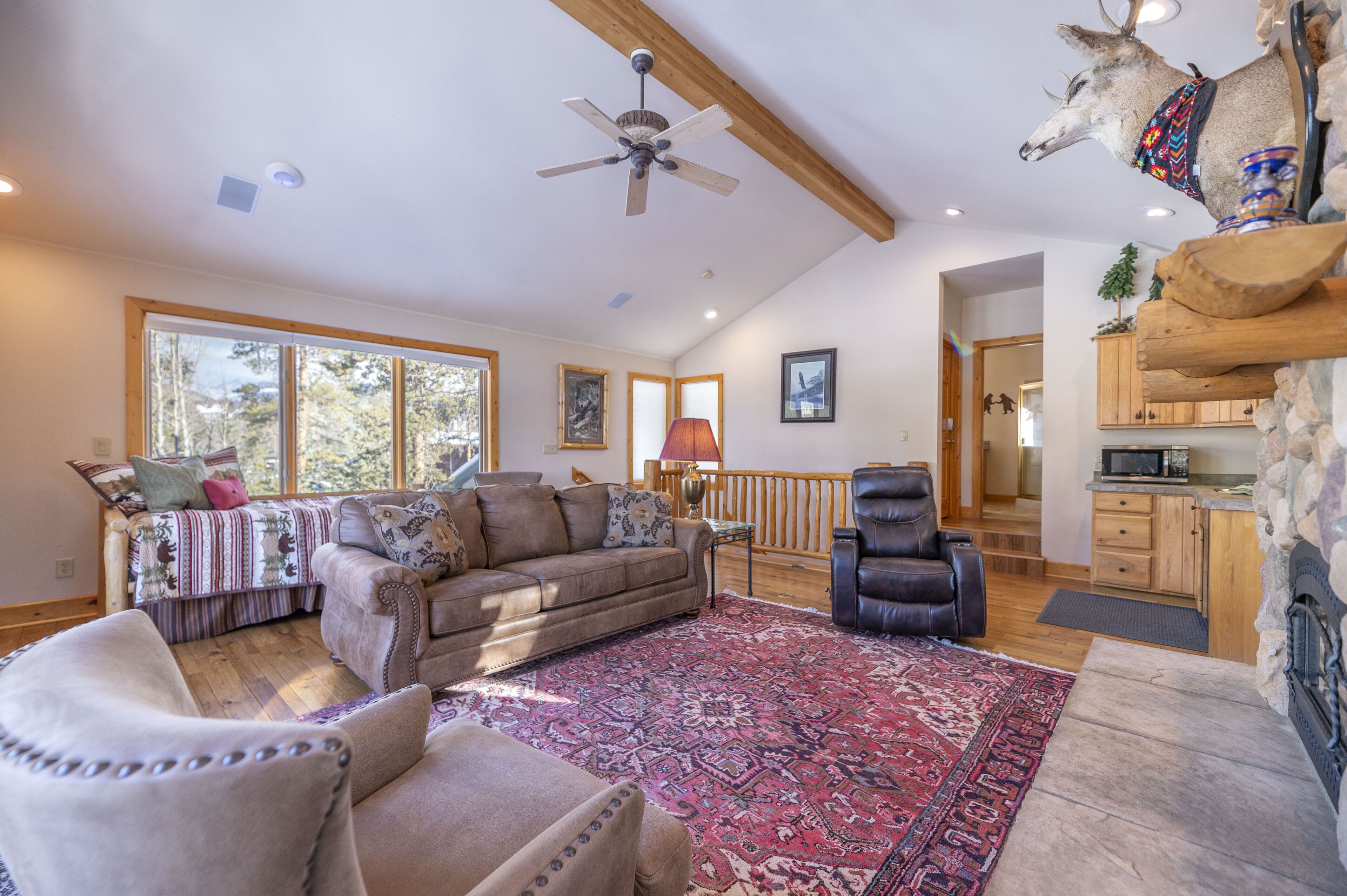 A spacious living room with a large window, a ceiling fan, a fireplace, and a kitchen area visible through an open door.